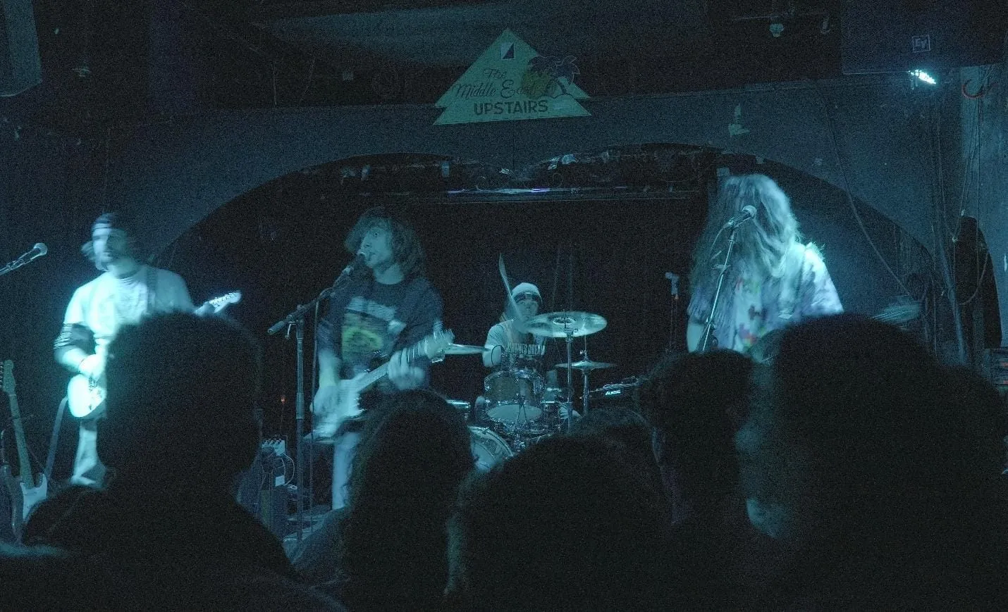 The image depicts a dimly lit indoor music venue with a band performing on stage, surrounded by a small audience.
AI-generated content may be incorrect.